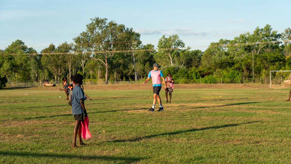 A young man plays football with a group of children. 