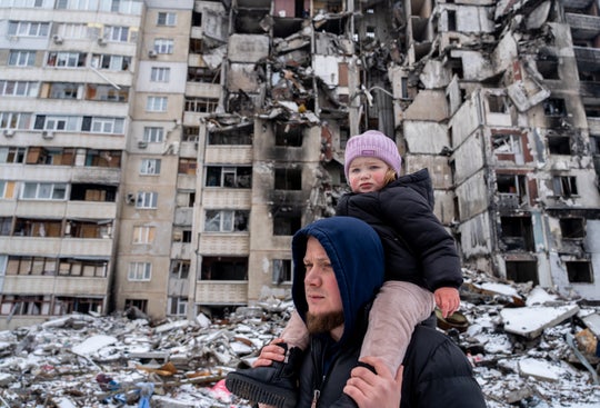 A father with his daughter on his shoulders standing outside a destroyed apartment building