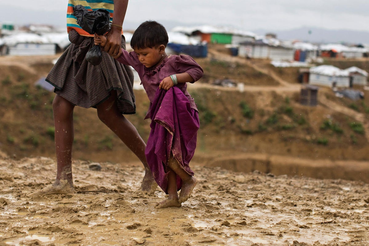 A young girl is walking barefoot on a muddy road. She is holding a woman's hand.