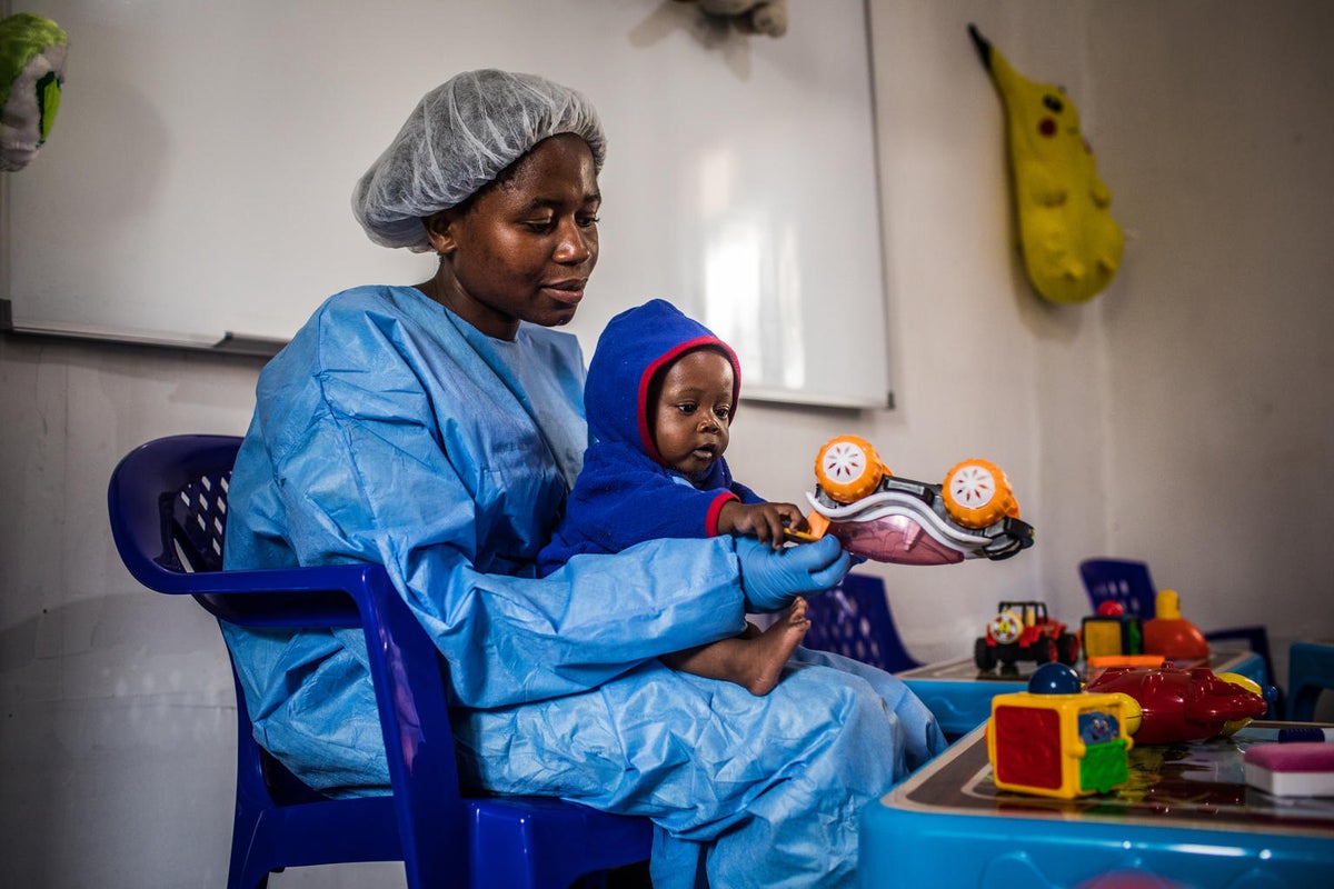 A woman wearing a protective gown, gloves and hair cover is playing with a toddler. They have a toy car.