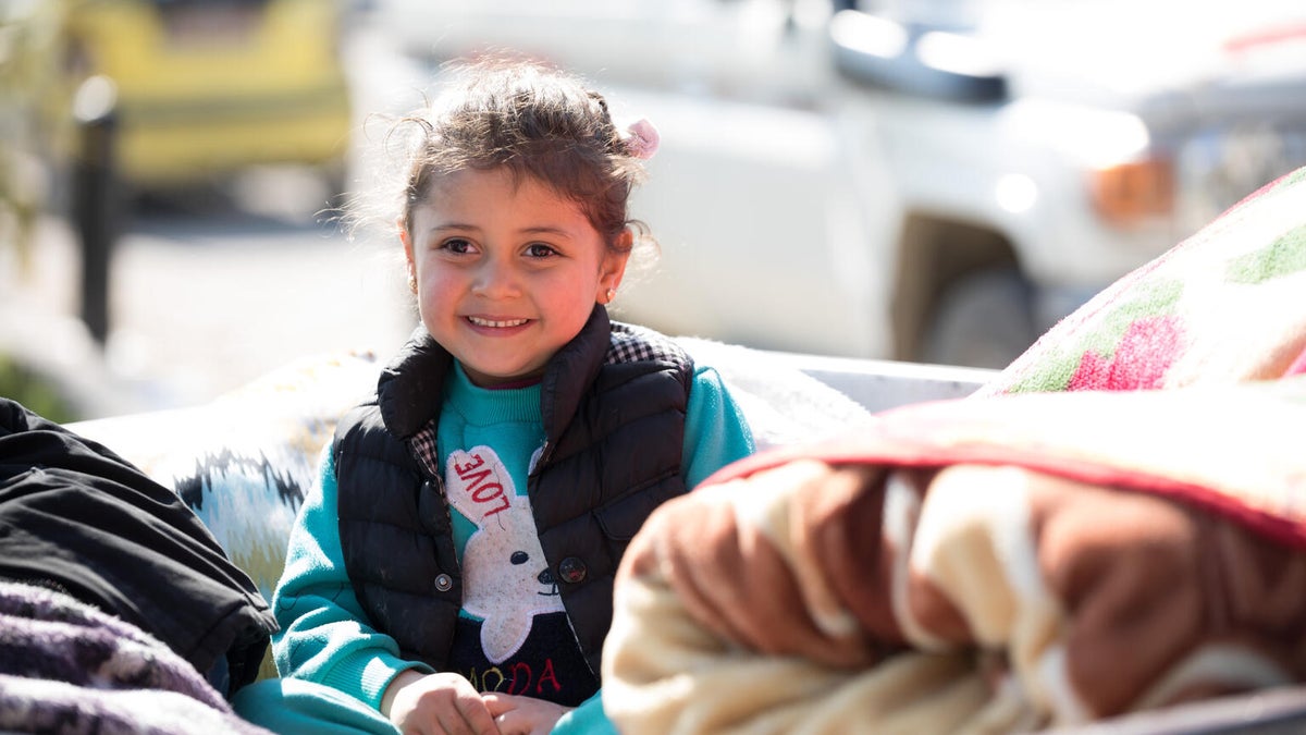 A Syrian girl smiles as she sits in a pile of blankets in the back of a car. 
