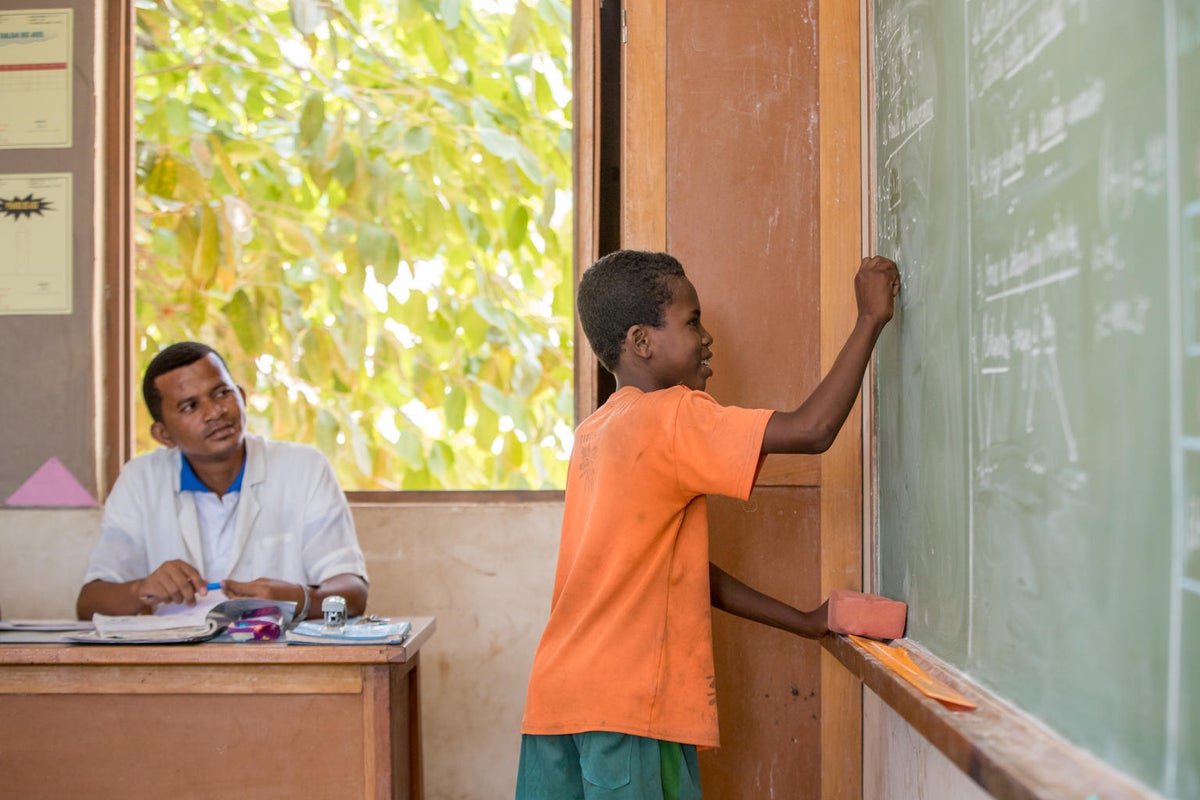 A boy is writing on a chalkboard at school. His teacher is looking at him.