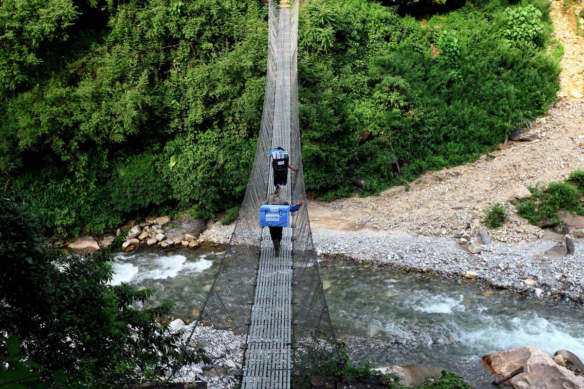 Two people walk over a suspension bridge carrying cooler boxes on their backs.