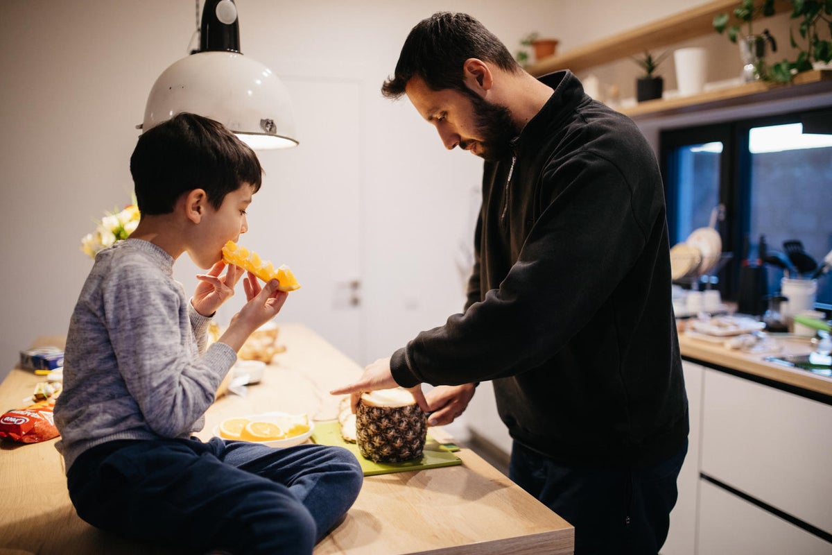 A man is peeling pineapple on a kitchen bench. A young boy is looking at him while we eats an orange.