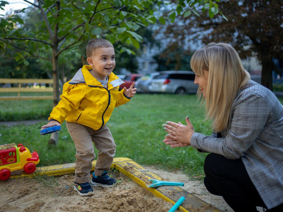 A young boy wearing a yellow parka smiles as he plays with a woman in a sandpit