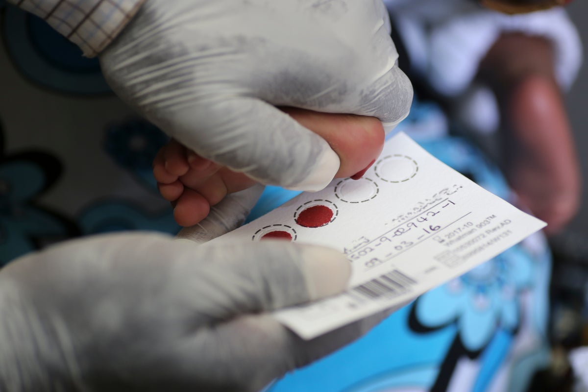 A pair of hands with gloves take a prick blood sample from a finger and deposit it on a paper card.