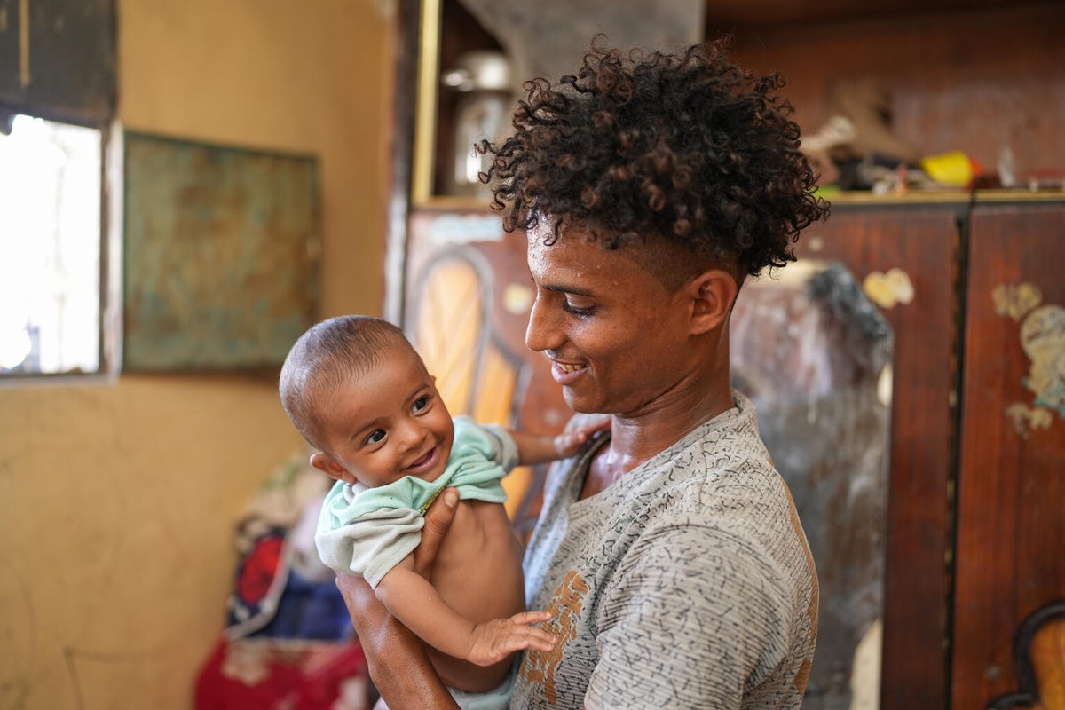 A young man smiles as he holds his infant child in his home in Yemen. 