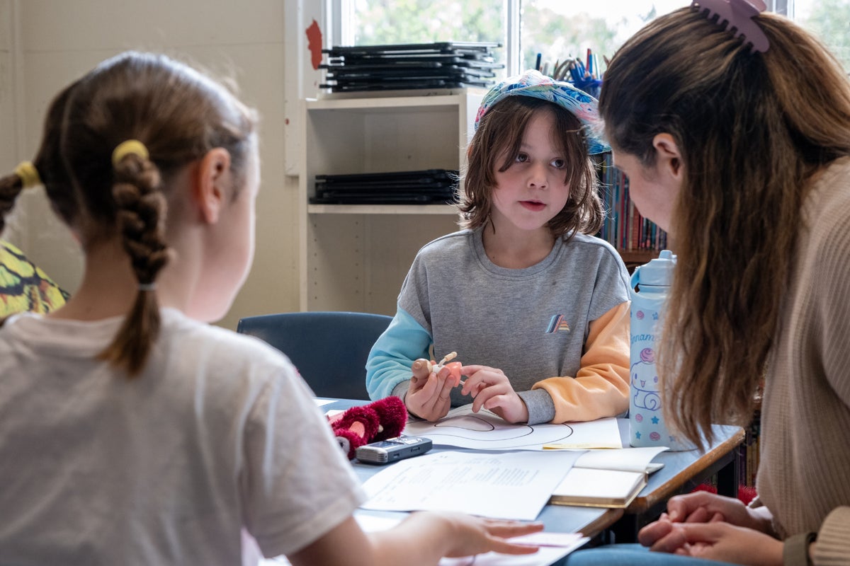 A young child sitting at a desk holding a figurine talks to a woman.
