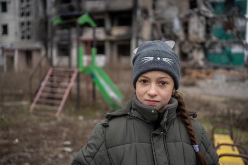 A girl looks at the camera in front of a pile of rubble that used to be her school.