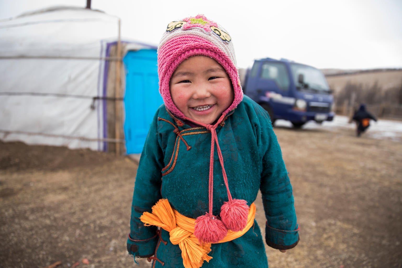 A young child smiles to the camera. He is wearing a beanie and a Mongolian garment.