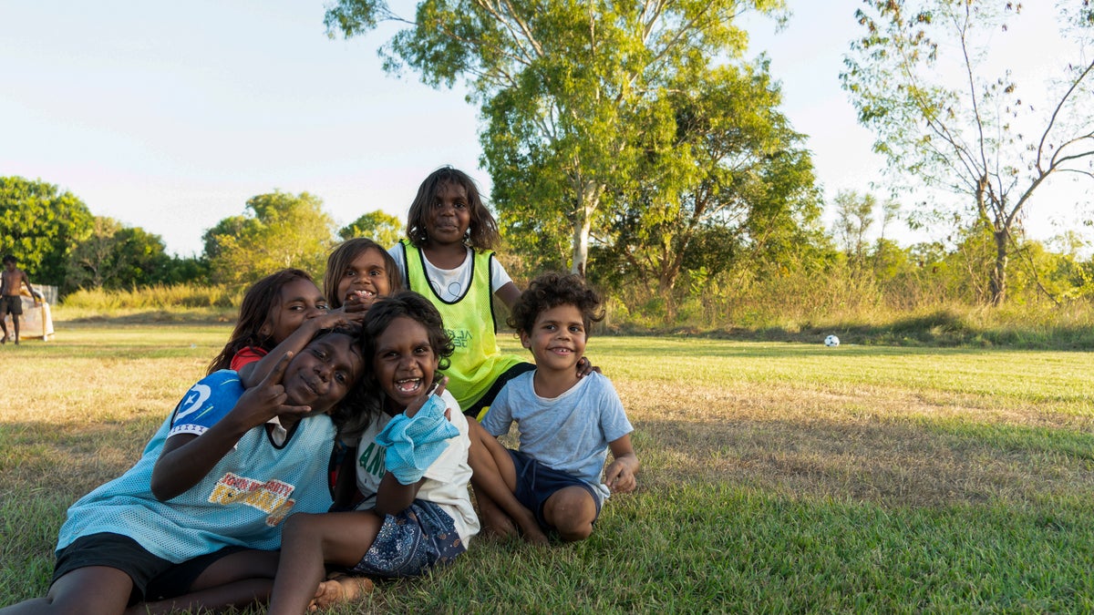A group of children smile sit on the grass and smile at the camera.