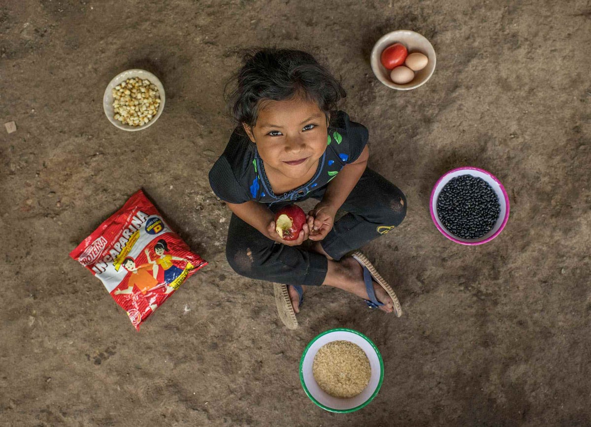 Jessica, fives, eats an apple and poses on the floor of the patio of her house. In the dishes there are beans, corn, rice, tomatoes, eggs, and Incaparina 