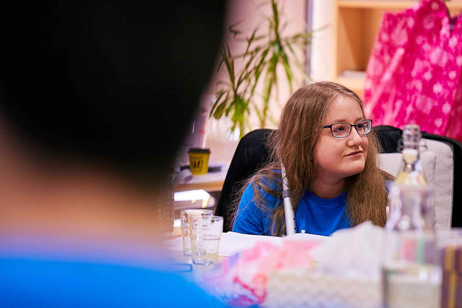 A young woman wearing a UNICEF t-shirt and glasses is sitting at a table hearing from other young people