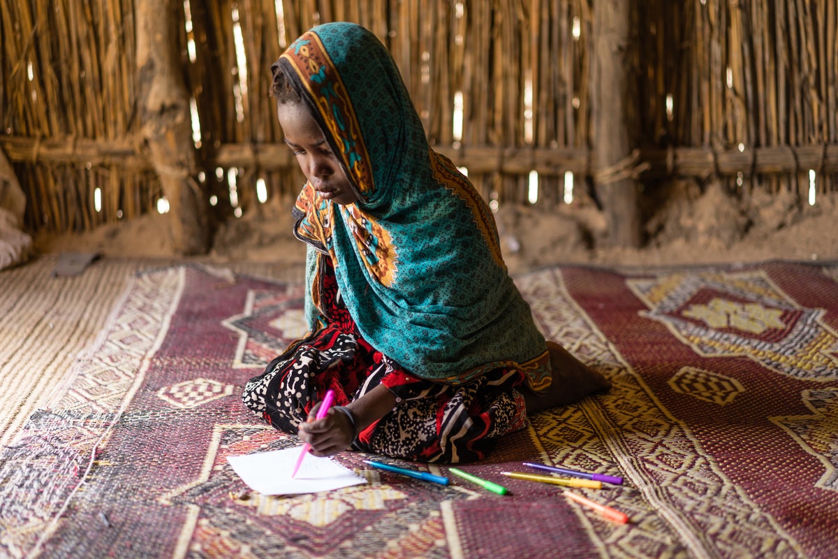 A young girl is doing homework on the floor inside a bamboo hut.