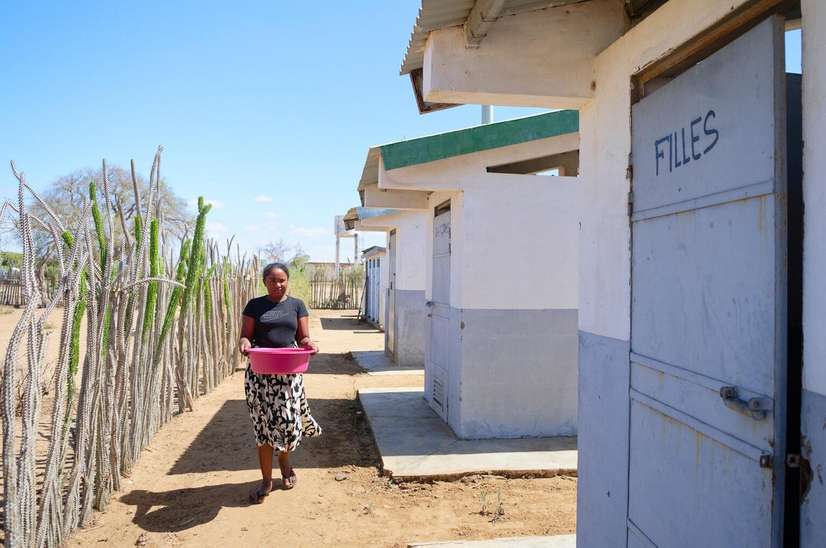 A teenage girl carrying a basin filled with water and heading towards a shower cubicle.