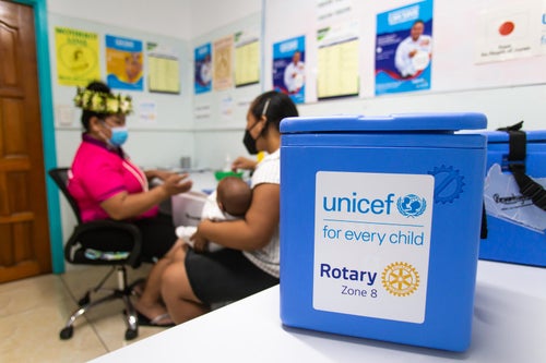 A blue UNICEF- and Rotary-branded vaccine storage box sits on a table in the foreground, while a nurse consults a mother and her infant child in a Cook Island medical centre in the background. 