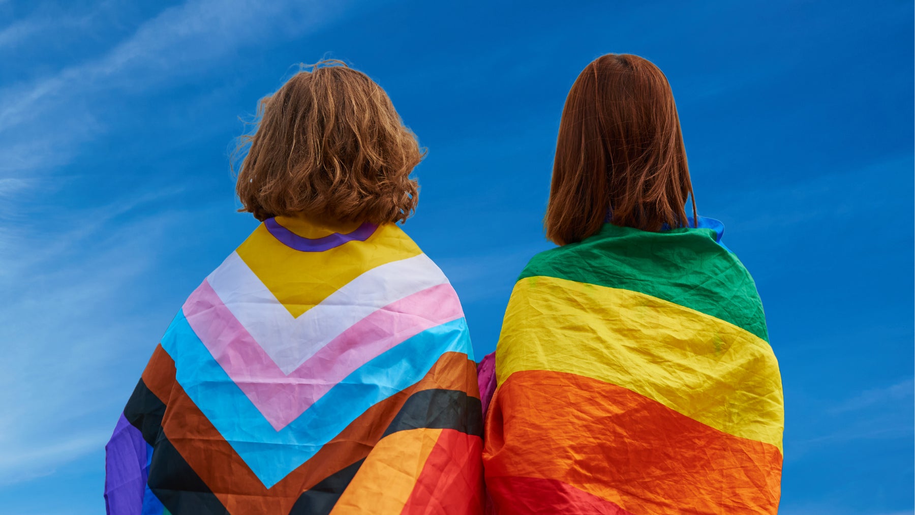 Young people wearing bright coloured LGBTIQA+ flags looking into sky