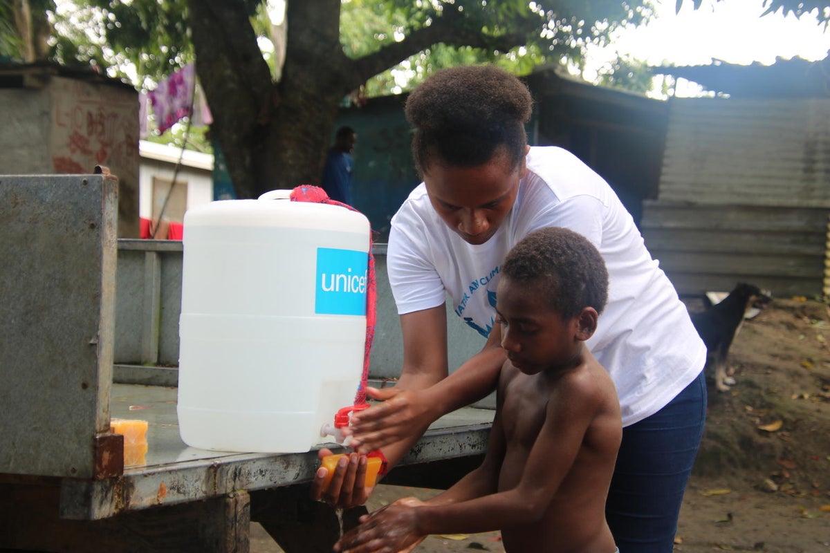 A woman is helping a boy wash his hands with water from a water dispenser. 