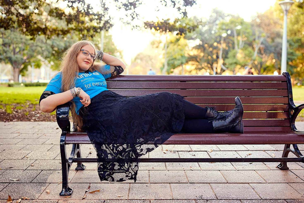 A young women wearing a UNICEF t-shirt and reading glasses is laying across a bench in a park