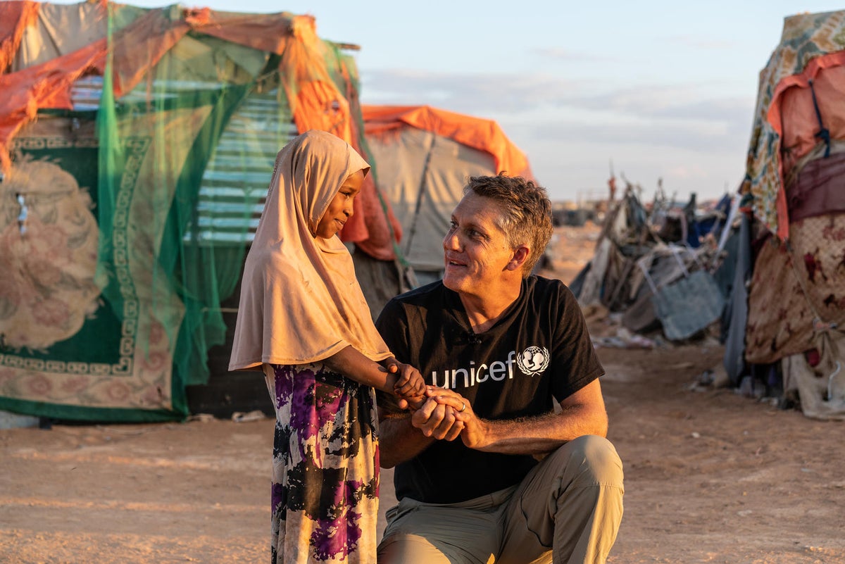 A man is squatting to be at the height of a young girl. They are talking. In the background, there is a displaced persons camp.