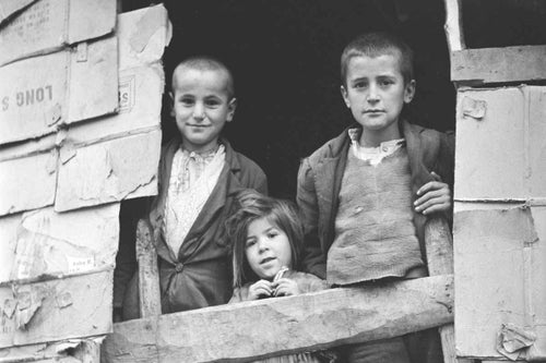 In Greece, 1949, refugee children peer out the window of a makeshift hut whose walls are made from cardboard boxes. 