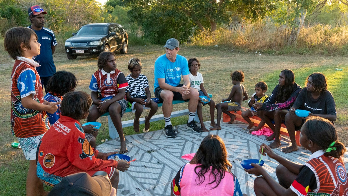 A group of children gather on benches and listen to a young man telling stories.