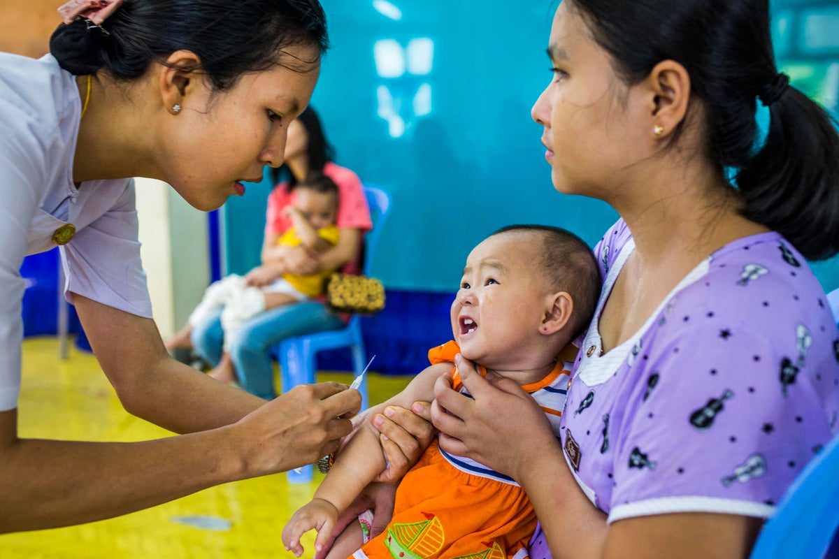 A baby is on her mum's lap while a health worker is about to apply a vaccine.