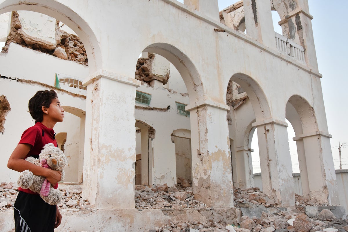 A boy holds a teddy bear while he looks at a destroyed house.