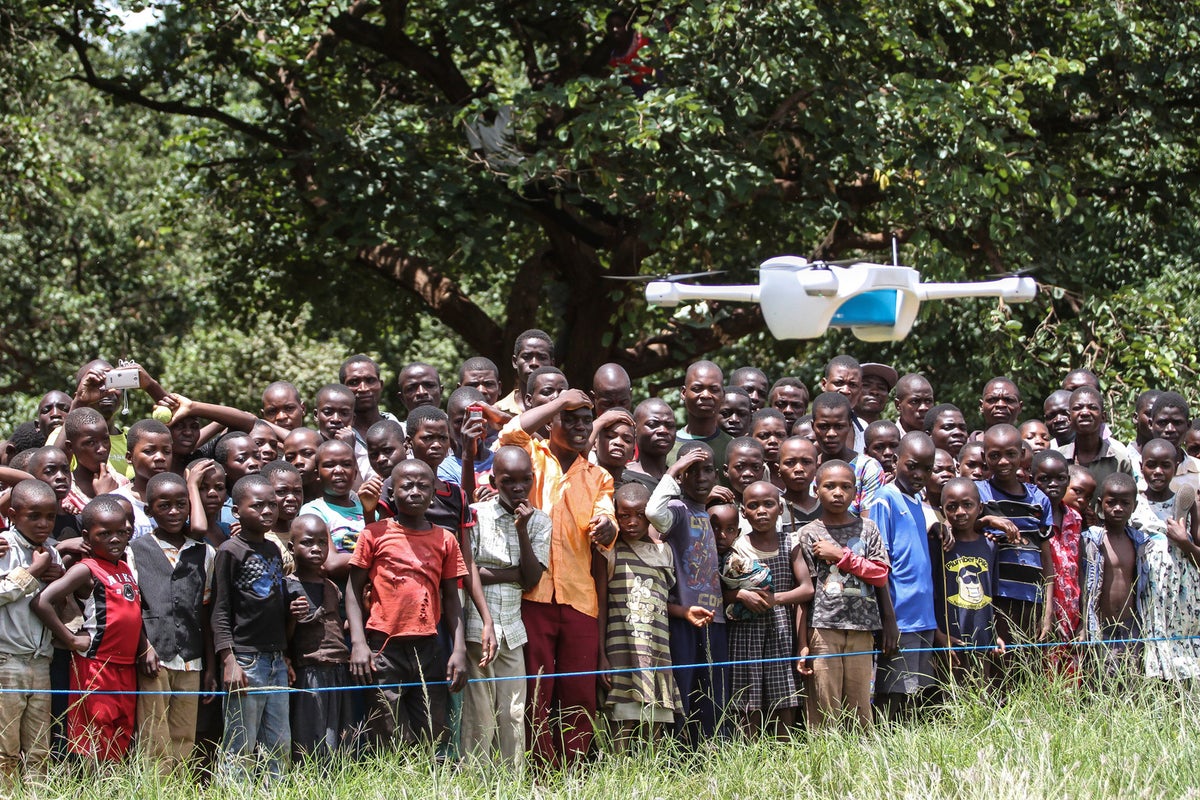 A big group of children look at a drone while it's landing.