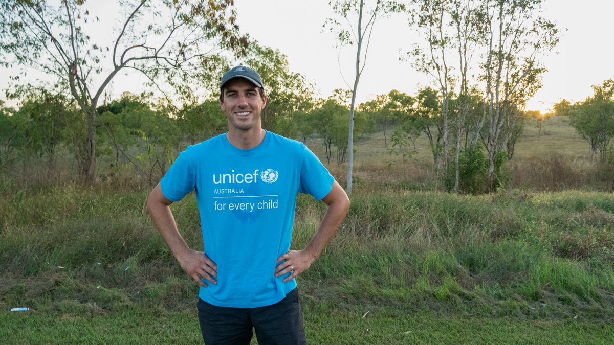 A young man smiles at the ground, standing on a grass field.
