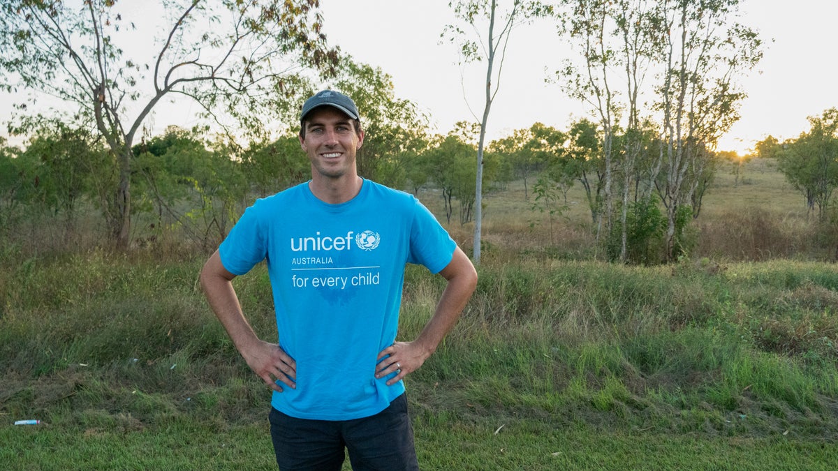 A young man smiles at the ground, standing on a grass field. 