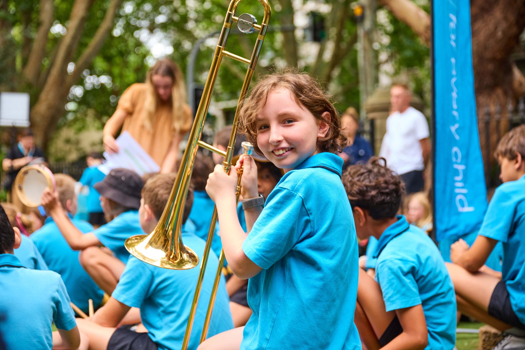 A student wearing a blue school uniform holds a trombone and smiles into the camera. 