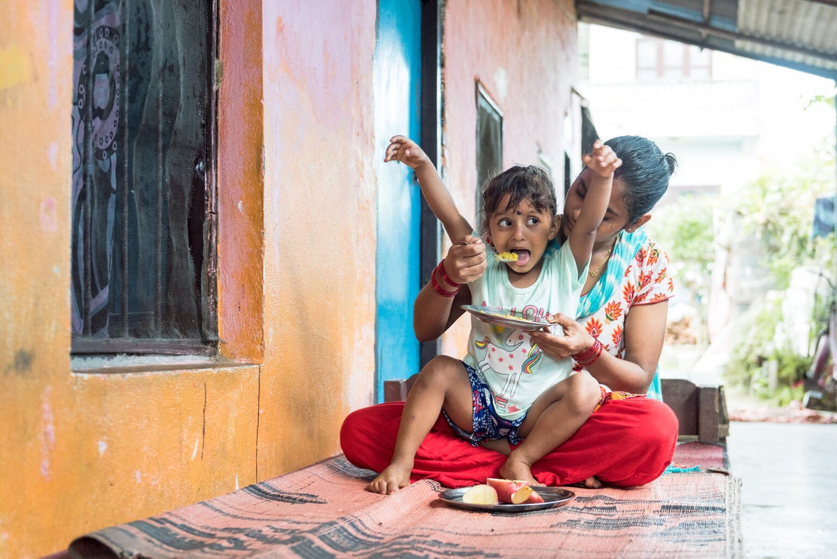 A young girl is seating on the lap of a woman. The woman is feeding the girl and the girl looks happy.