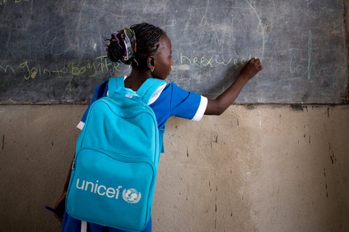 A girl writing on a chalkboard. She is wearing a UNICEF backpack