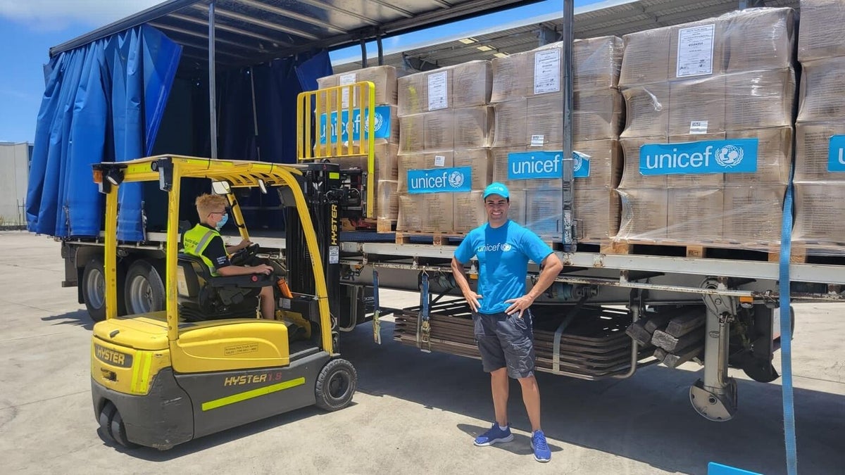 A forklift is loading pallets of supplies onto a truck. A man with a UNICEF shirt smiles in front of the truck.