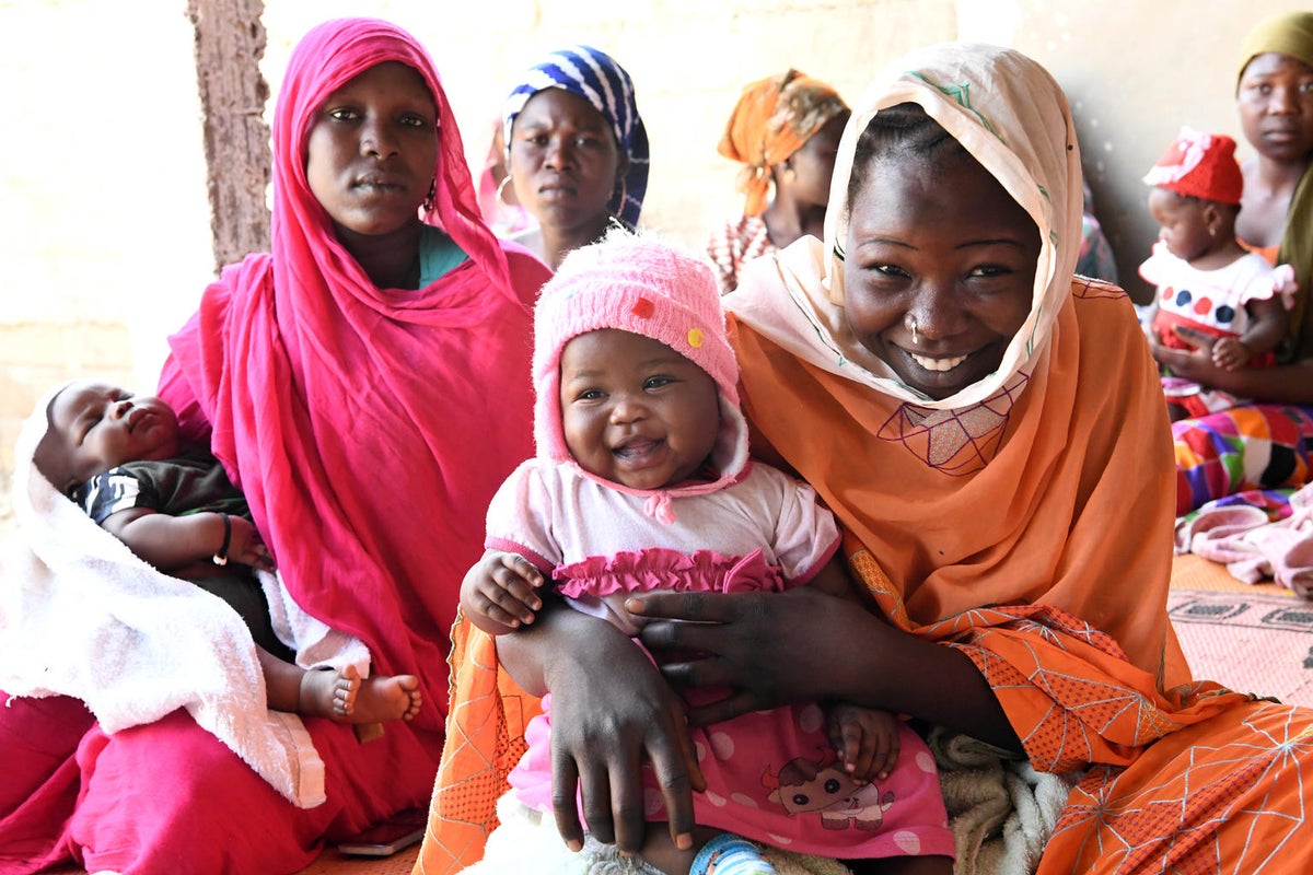 A woman is holding a baby girl on her lap and they are both smiling. Behind them, there's other women holding babies as well.