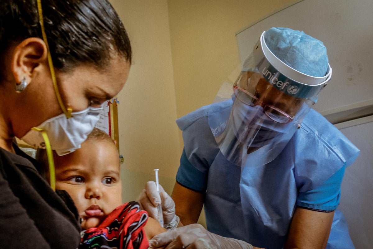 A baby held in the arms of his mother is receiving a vaccine in the arm provided by a health worker.