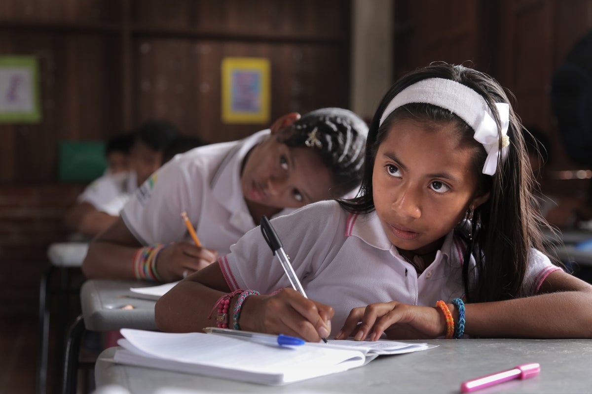 A girl is seating on a school bench and is writing something. She looks like she is paying attention to the teacher.