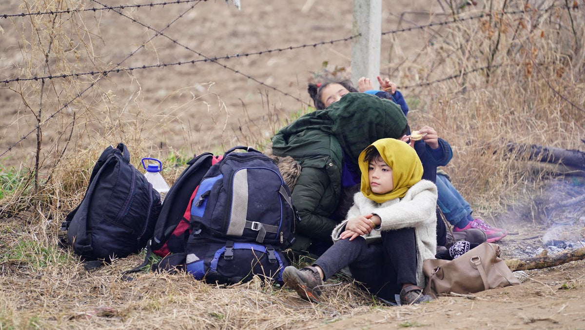 A group of children are seating on the floor by a barbed-wire fence. They have backpacks with them.
