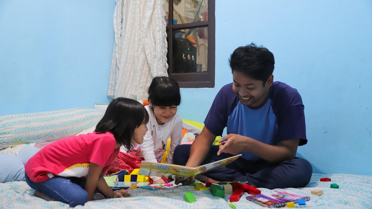A man is reading a book to two young girls. They are all smiling.