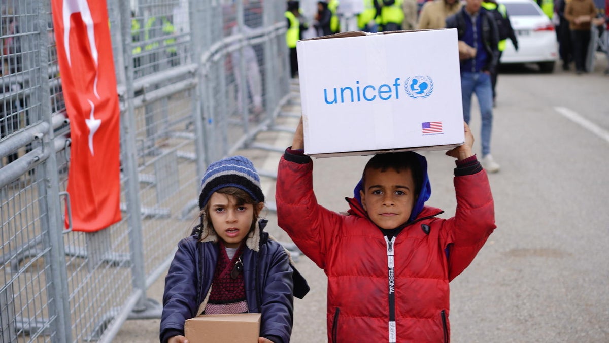 Two children walking down a street. One of them is carrying a UNICEF box over his head.