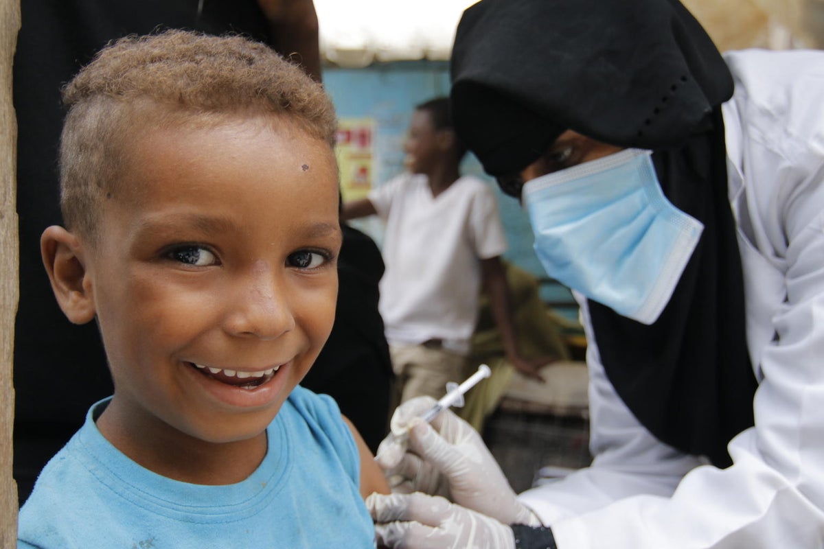 A boy is about to get a vaccine on his arm. He is smiling to the camera.