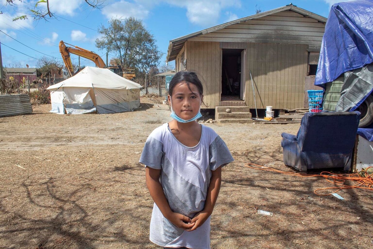 A girls poses to the camera. Behind her there's a house, and next to it, and emergency tent.