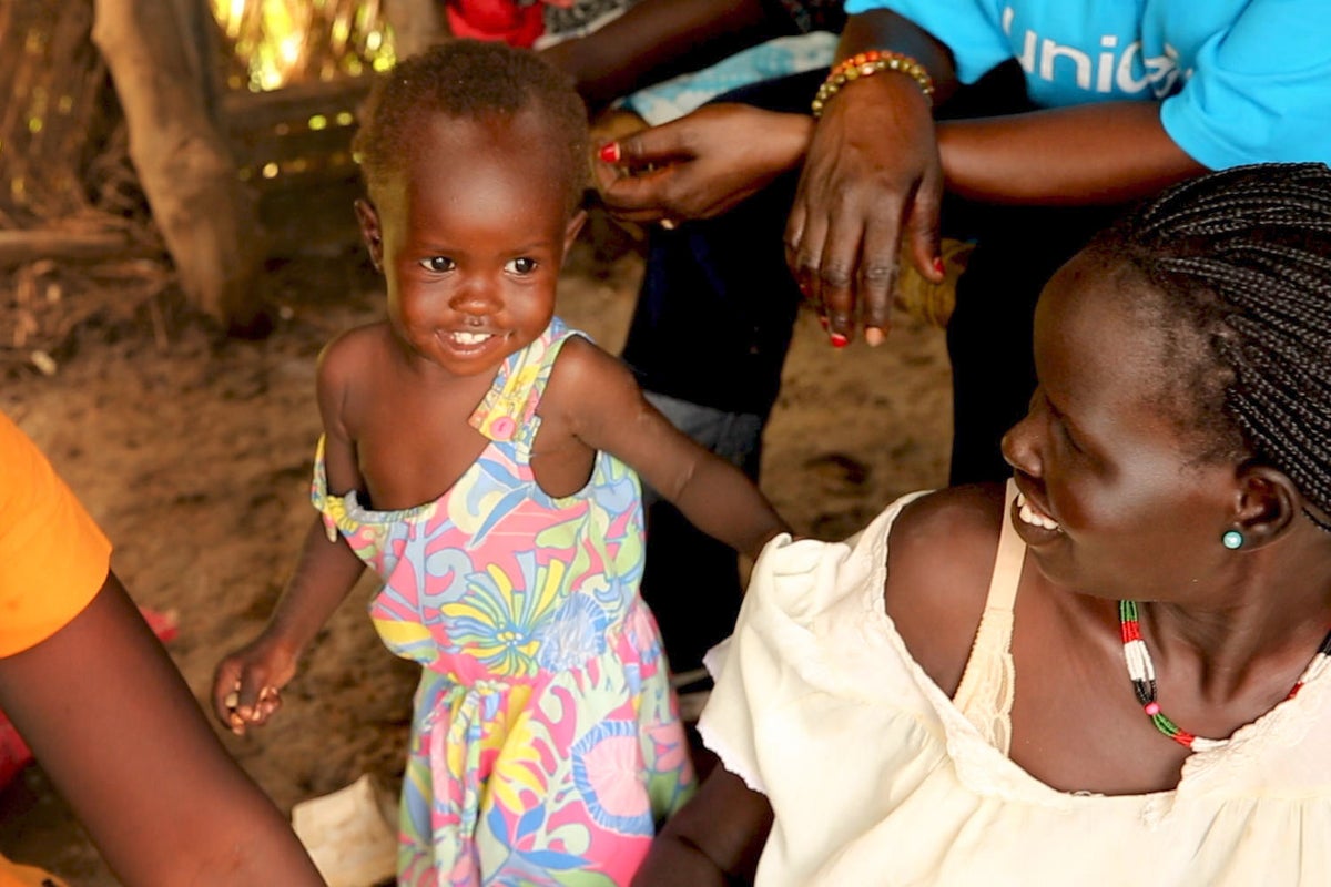 The toddler is looking happy and is standing next to her mother and a UNICEF worker