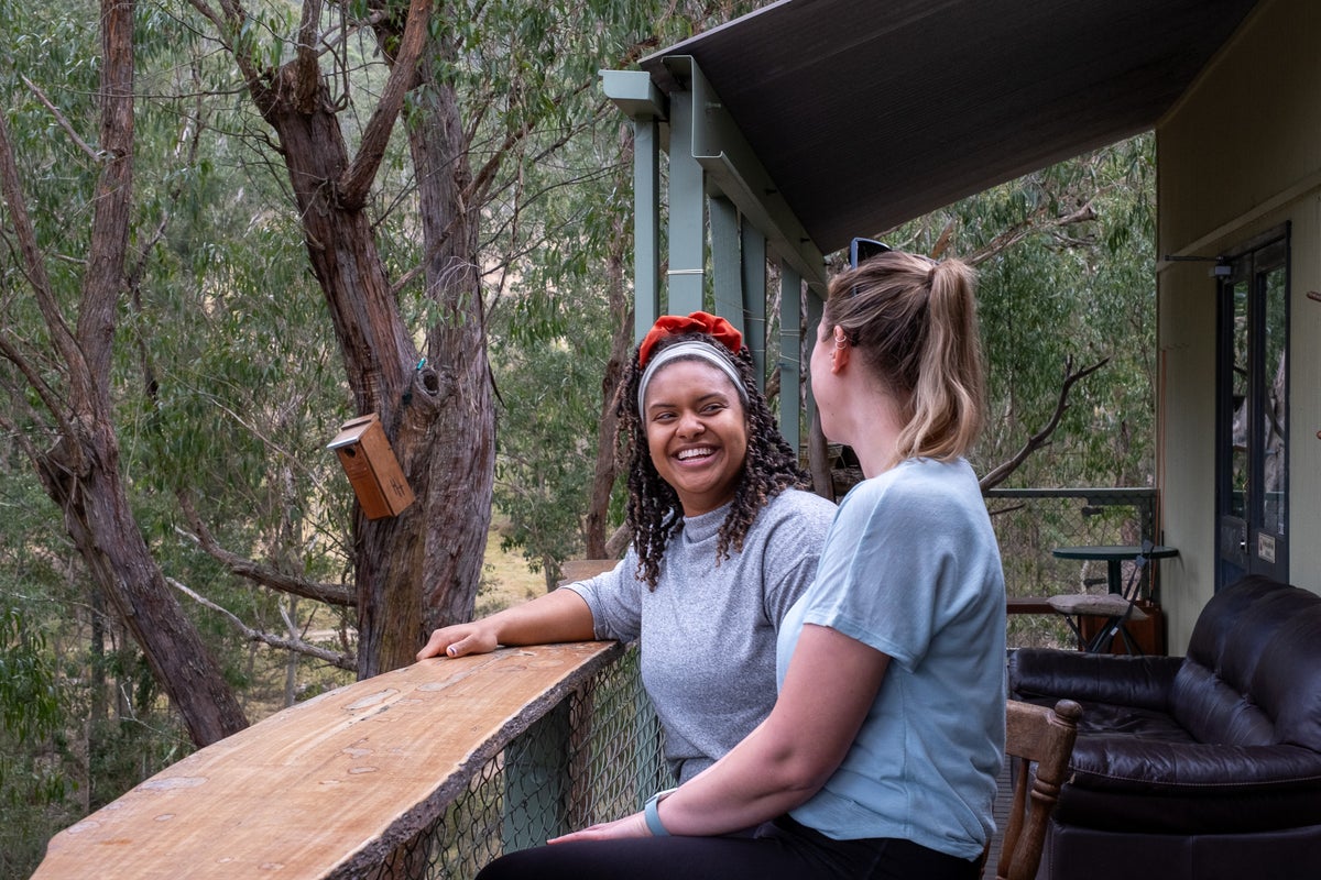 A young woman laughing in conversation with her female friend on a balcony surrounded by trees.