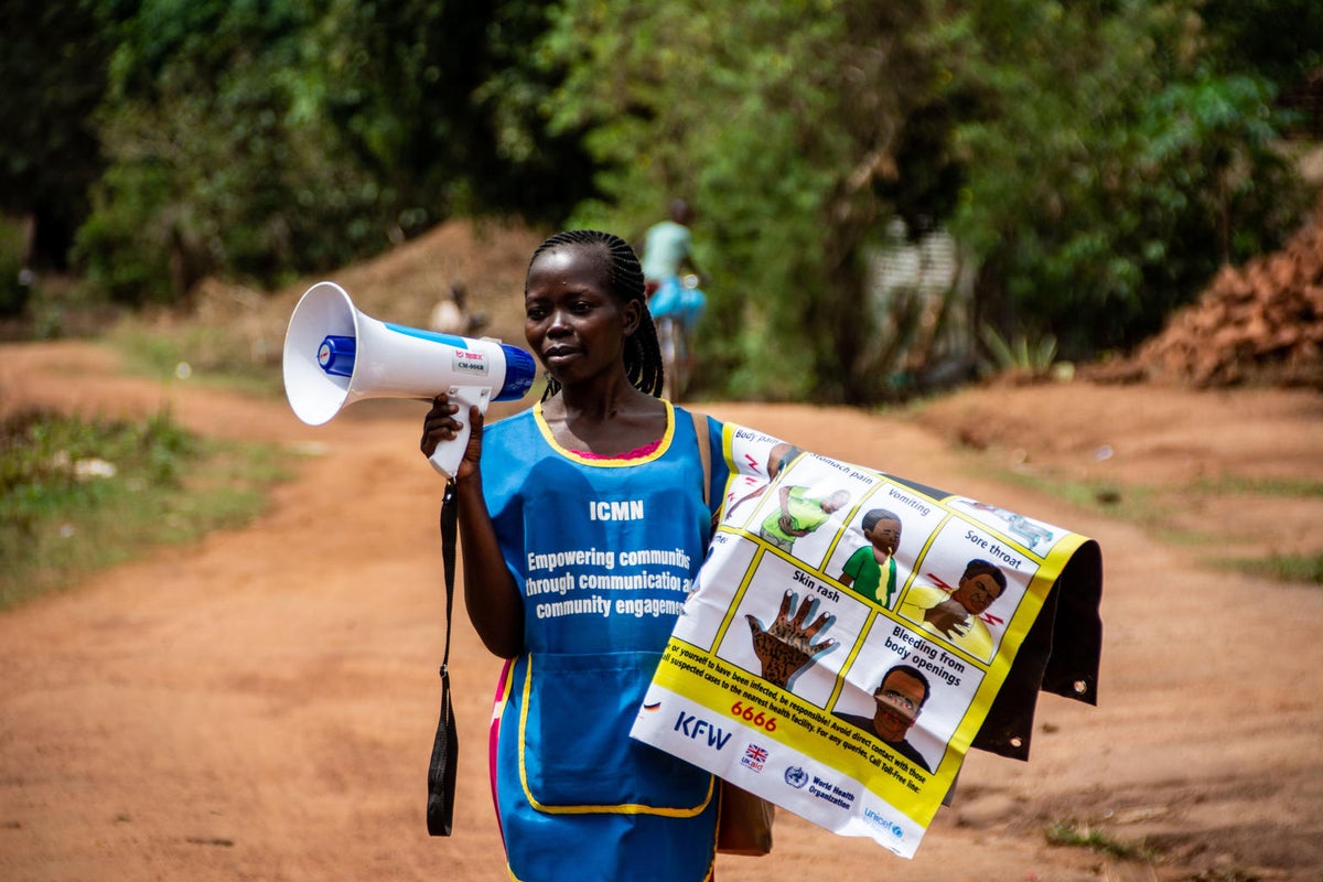 A woman is speaking through a megaphone. She is holding a poster explaining COVID-19 symptoms.