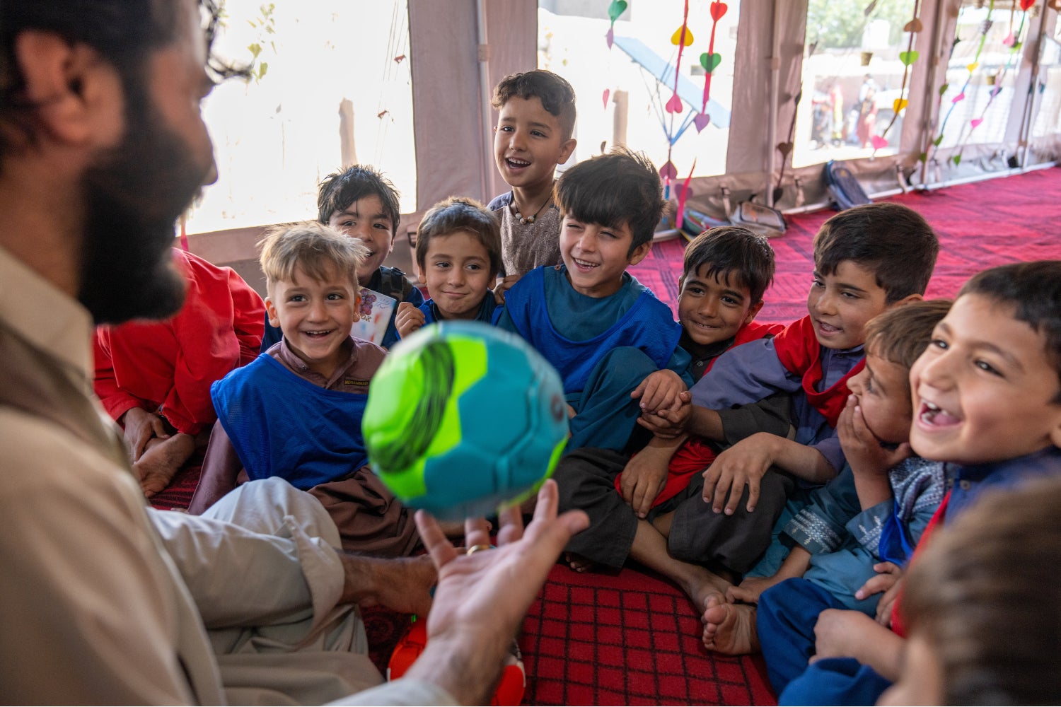 A group of young Afghani boys smile as they watch a man demonstate a ball game in a child-friendly space in Afghanistan.