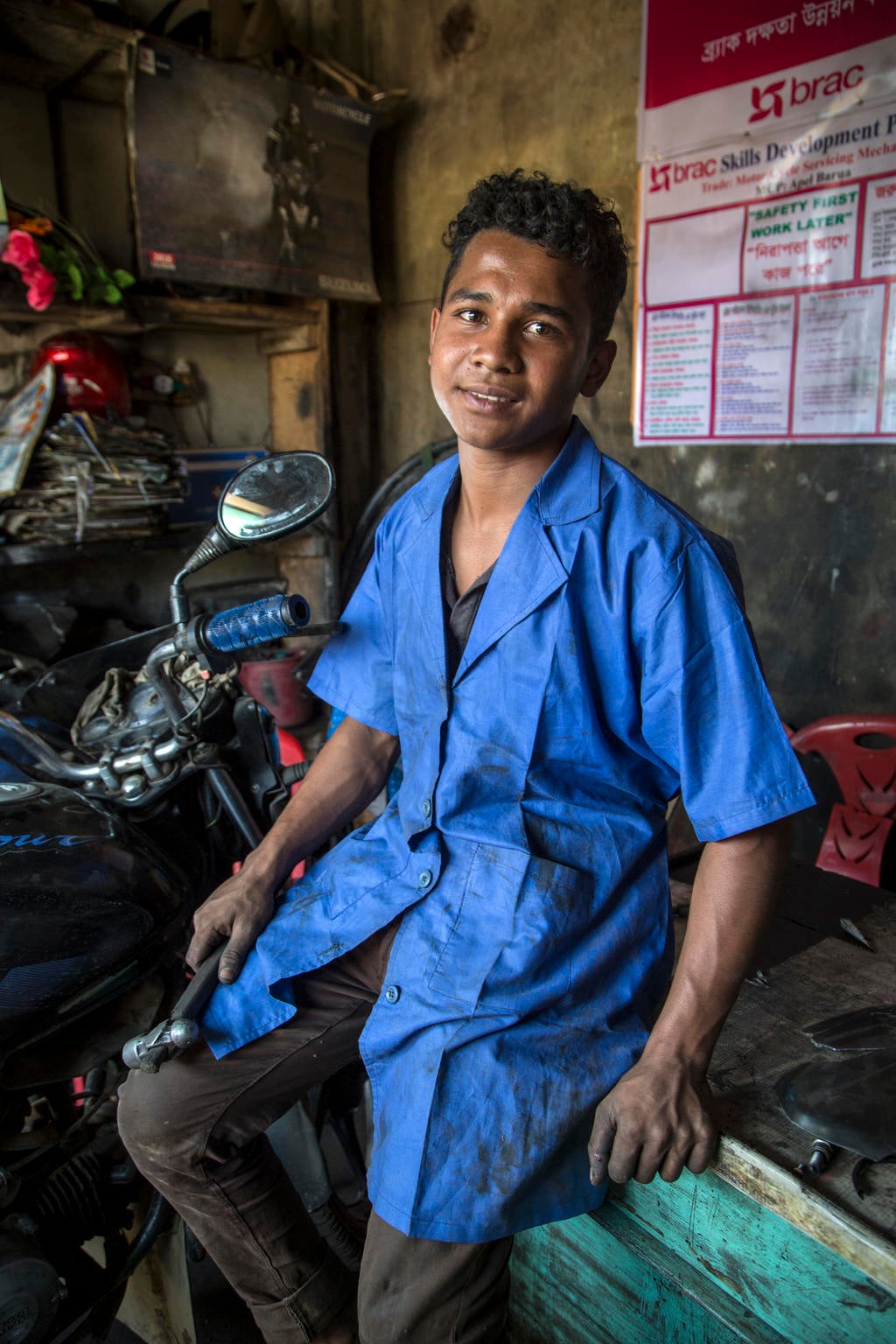 A young man smiles to a camera next to a motorbike. It looks like he is in a workshop.