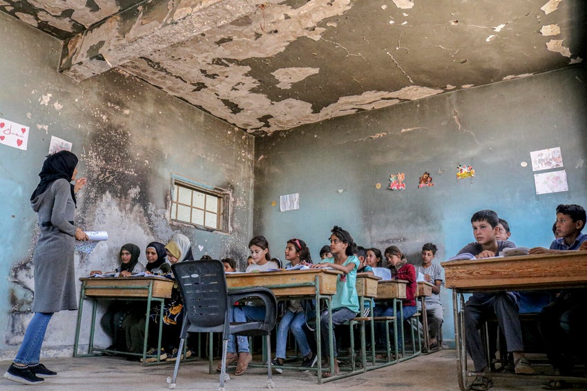 Students sitting at their desks in a damaged classroom, learning from a teacher standing in front of them.
