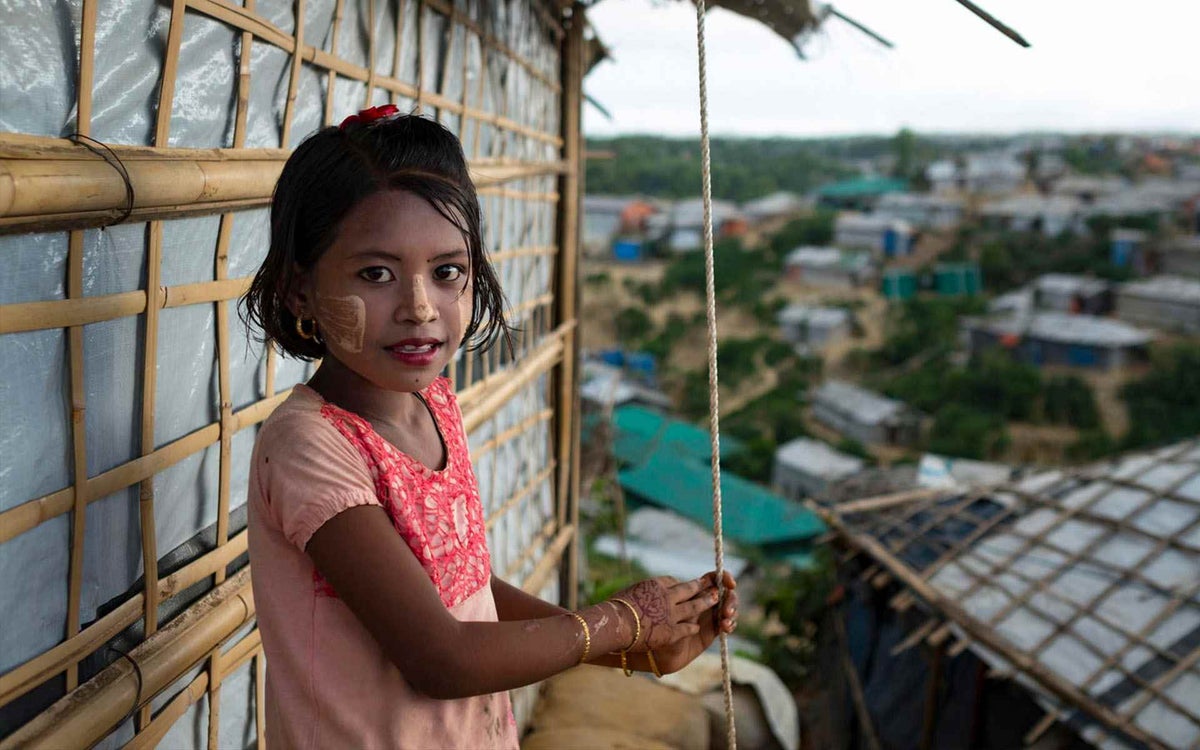 Sara, 7, stands against her family’s plastic-and-bamboo house in the Kutupalong refugee camp in Cox’s Bazar, Bangladesh